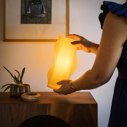 Terra Labs Woogie table lamp featuring a honey-colored rounded ceramic base and a white fabric lampshade on a wooden surface.